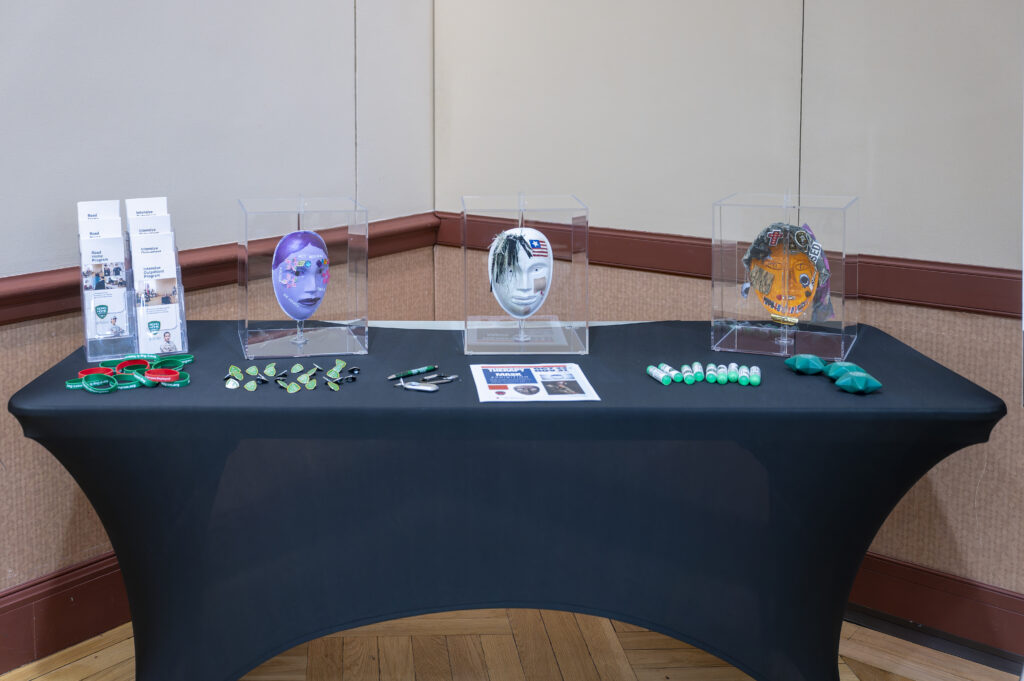 Three masks designed by veterans experiencing PTSD displayed in individual cases on a black tablecloth. The first mask, purple with gems, flowers, and words 'be happy' and 'act normal.' The second mask, white with a flag, bandages, hair, and a red cross. The third mask, orange with camo, patches, a cross, and cut-out words. The masks are accompanied by Road Home Program swag items
