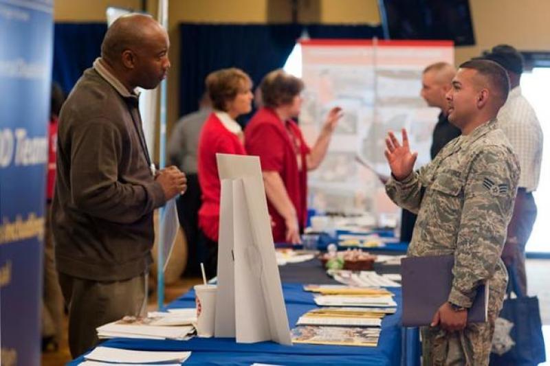 Man in military uniform speaking to man in civilian attire with a table/booth between the two.  In the background are other people talking in similar, career fair, type style with tables/booths sharing organization information/pamphlets. 