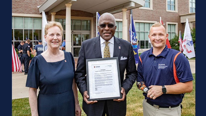 Dean Cheryl Hanley Maxwell stands with Chancellor Jones and Garrett Anderson in front of the Chez Veterans Center, holding the designation of a Purple Heart University in a frame. 