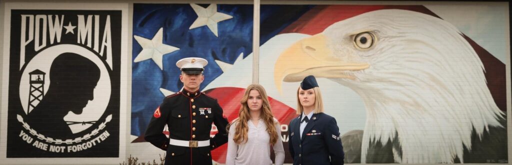 Three siblings stand in front of a POW memorial with a flag and eagle painted on a brick wall.  The siblings stand shoulder to shoulder.  On the left, is the tallest and only male, wearing and US Marine Corp uniform.  in the middle, is a woman in a white long sleeve shirt with curly long light brown hair.  On the right is a woman with blond hair wearing an air force uniform. 