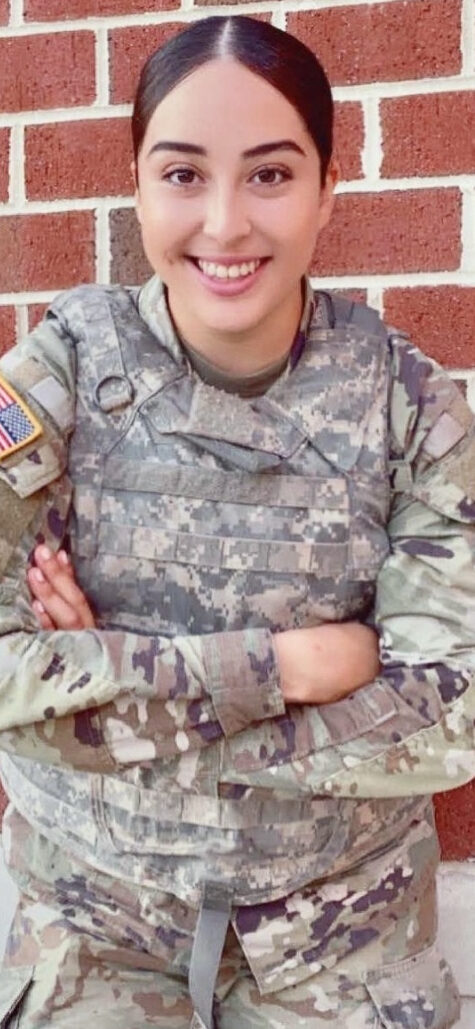 A woman stands with her arms crossed wearing a army uniform.  She is smiling and looking towards the camera.  Her hair is dark and pulled back away from her shoulders and face. She stands against a brick wall. 