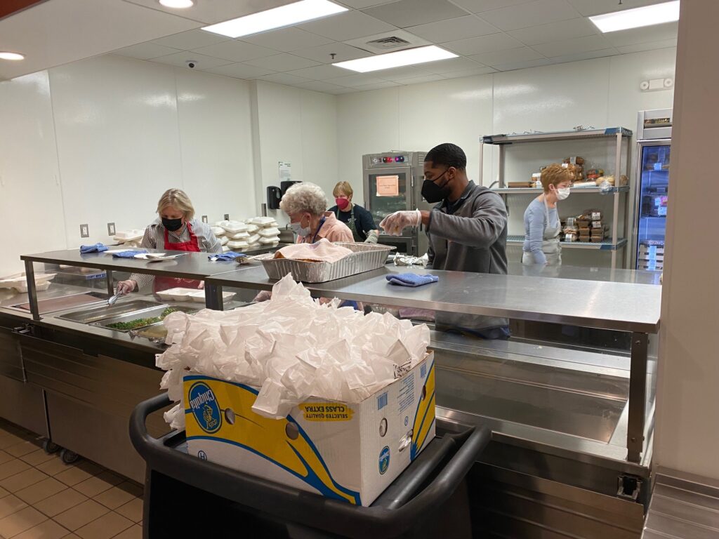 Four individuals stand behind a metal counter in a kitchen.  Three of the individuals are older women, one is a black man.  All appear to be working on prepping meals into containers. 