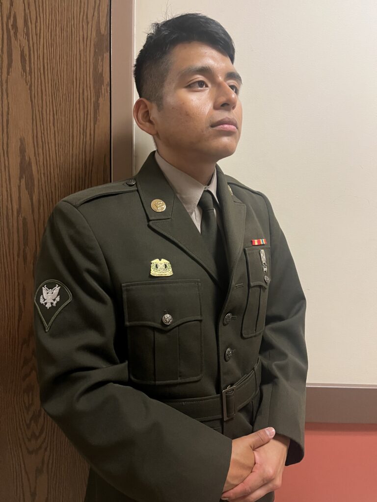 Cristian is standing in front of a wooden door staring away from the camera.  Cristian is wearing his Army Service Uniform. 