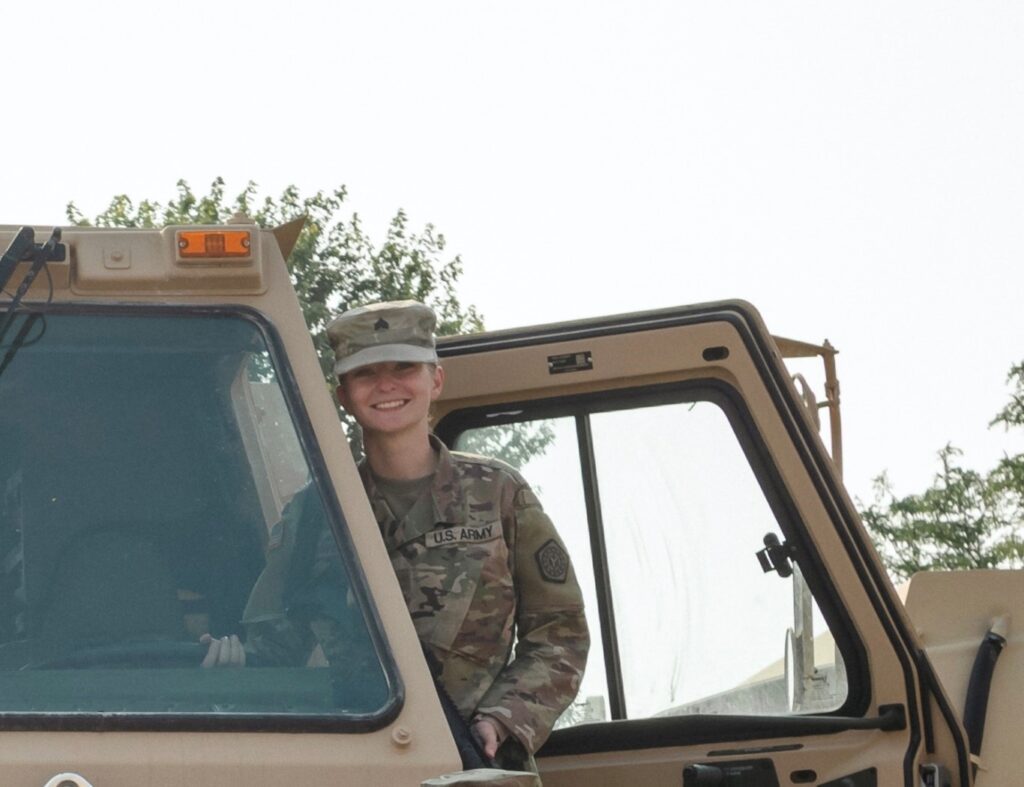 Cloie peeks out of a Humvee in her Army uniform.  She is smiling and looking directly at the camera. 